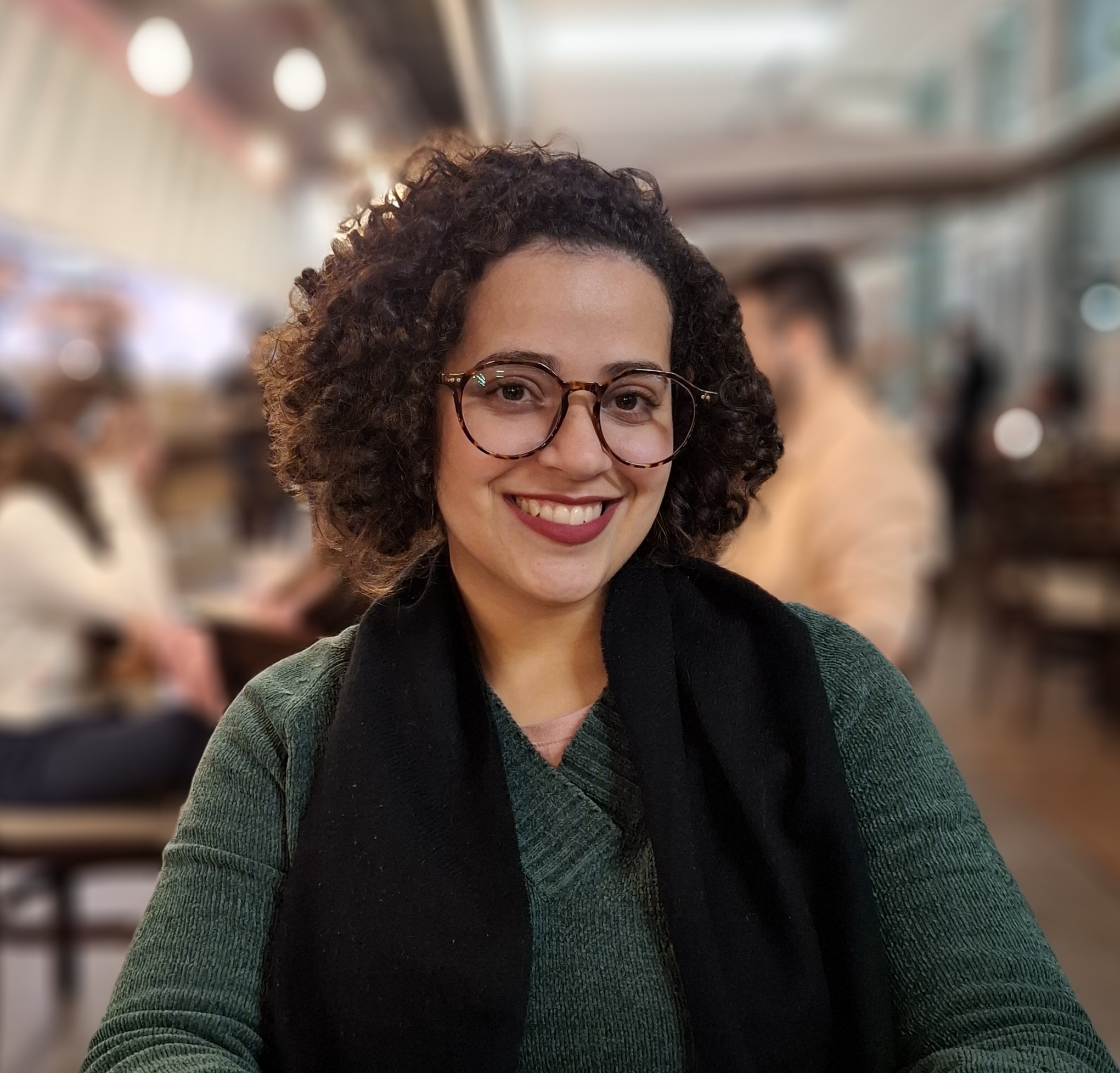 Retrato de uma mulher branca de cabelo castanho cacheado na altura do pescoço, óculos arredondado, batom vinho e suéter verde escuro com cachecól preto.
